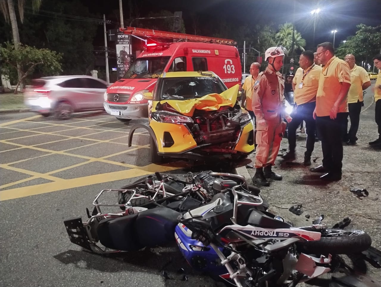 Acidente envolvendo moto e táxi no bairro da Lagoa, no Rio de Janeiro, com motocicleta destruída no chão, táxi com a frente totalmente danificada e equipe do Corpo de Bombeiros realizando atendimento no local.